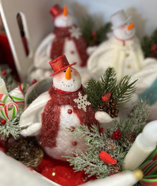 Little white snowman made of fabric, with red knitted scarf and red hat, keeping some Christmas decorations in hand